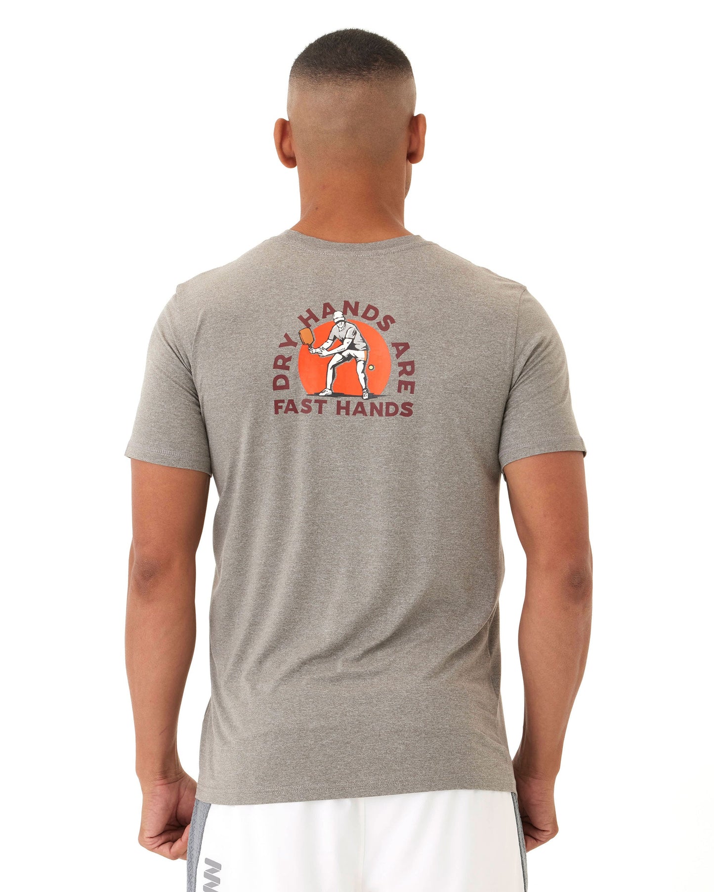 A person faces away wearing the Wringer Wear Men's Short Sleeve Fast Hands Graphic Tee in light gray, featuring “DRY HANDS ARE FAST HANDS” in red above chalky hands, paired with white shorts. The athletic cut offers a performance-ready look.