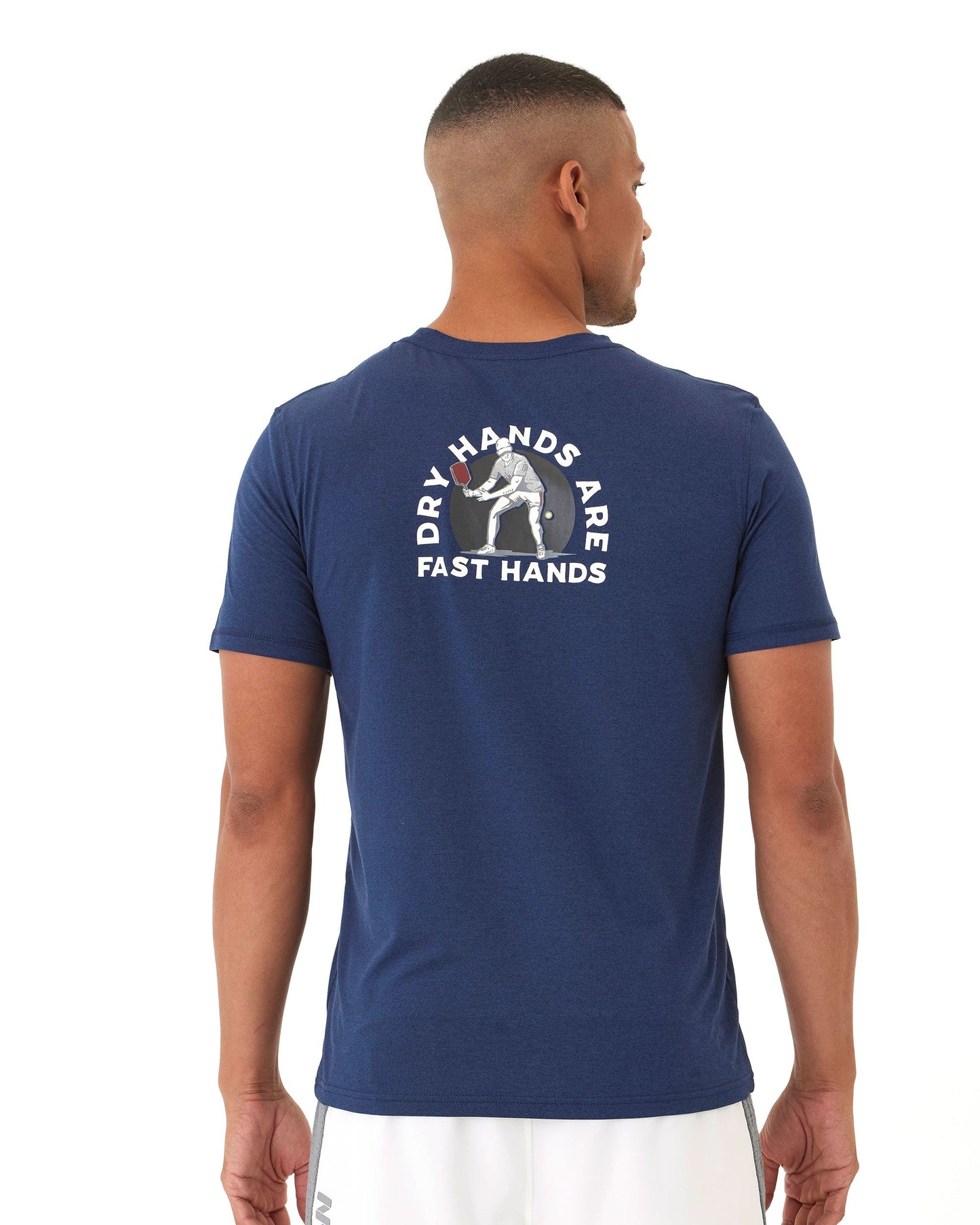 A man with short hair, seen from behind, models the Wringer Wear Men's Short Sleeve Fast Hands Graphic T, featuring “DRY HANDS ARE FAST HANDS” and an illustration on the back. He pairs it with white shorts against a plain white background.