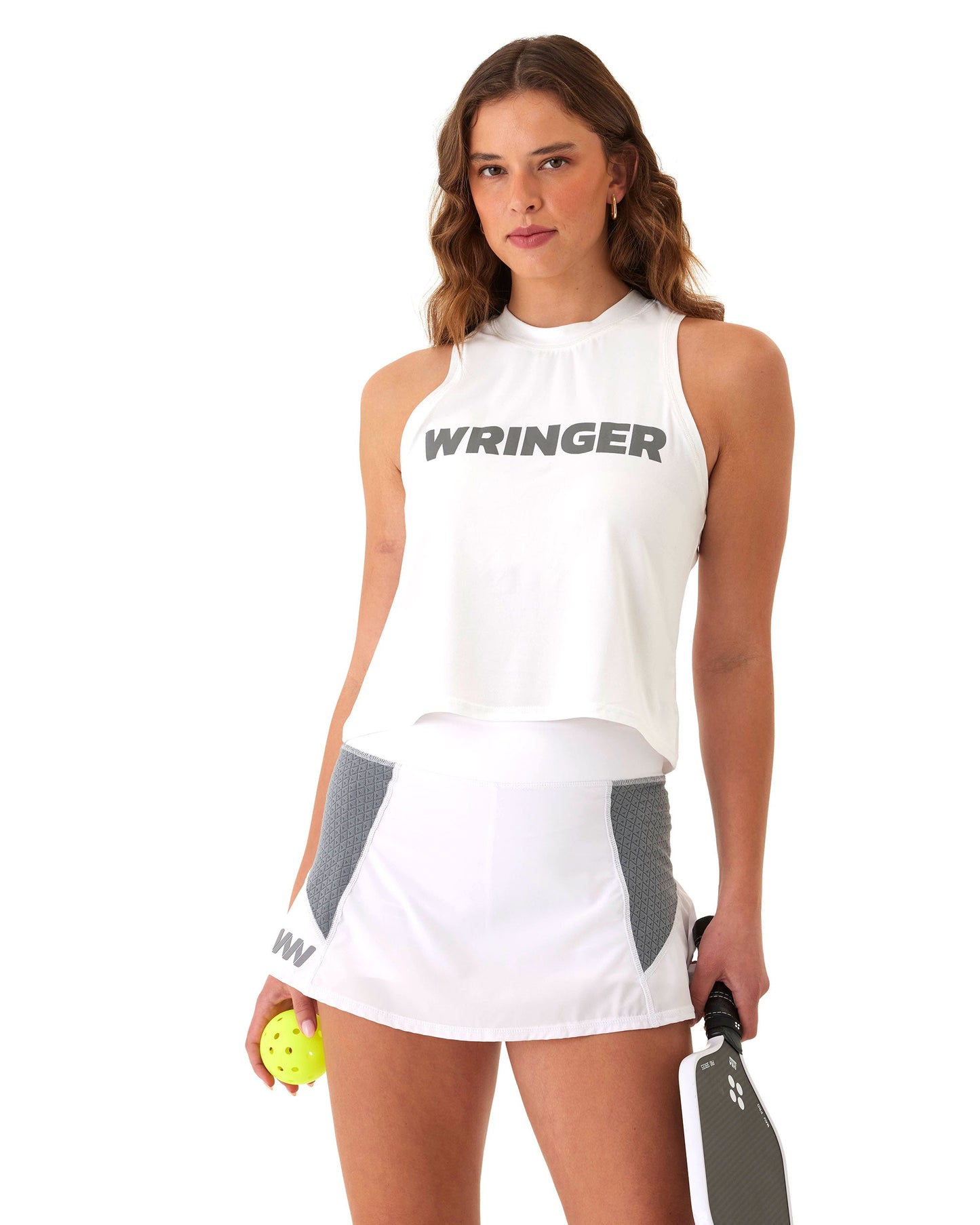 A woman with wavy brown hair wears a white sleeveless top and the WRINGER WEAR Moab Skirt, a moisture-wicking tennis skirt with gray panels. She holds a yellow pickleball and a black paddle, standing against a plain white background.