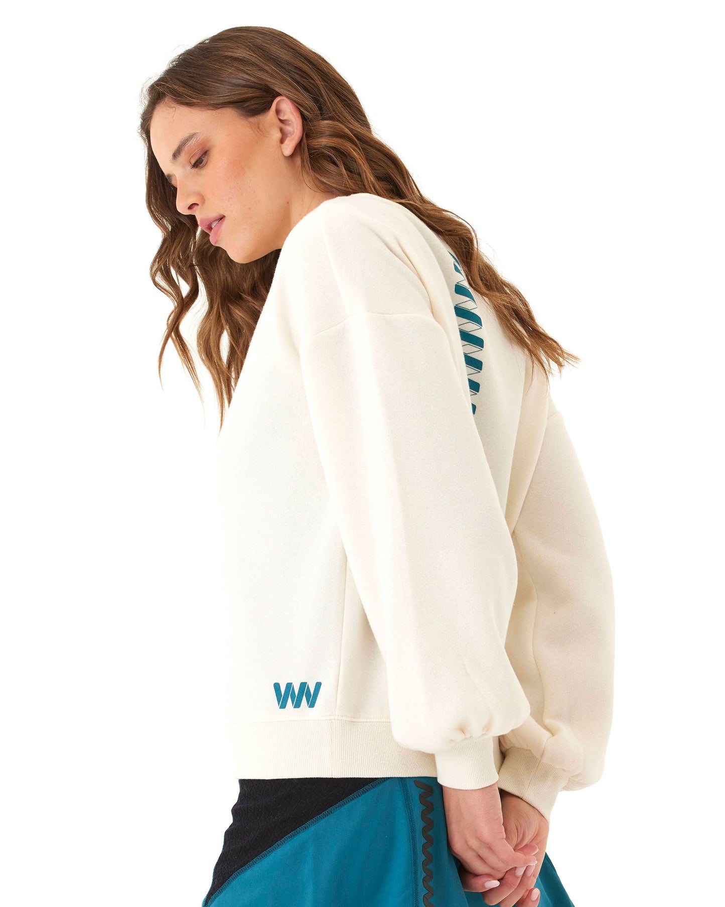 A woman with wavy brown hair models the Wringer Wear Women's Crew Neck Sweatshirt, cream-colored with blue WW initials near the hem and vertical blue text on the back. She pairs it with a teal and black skirt against a white background.