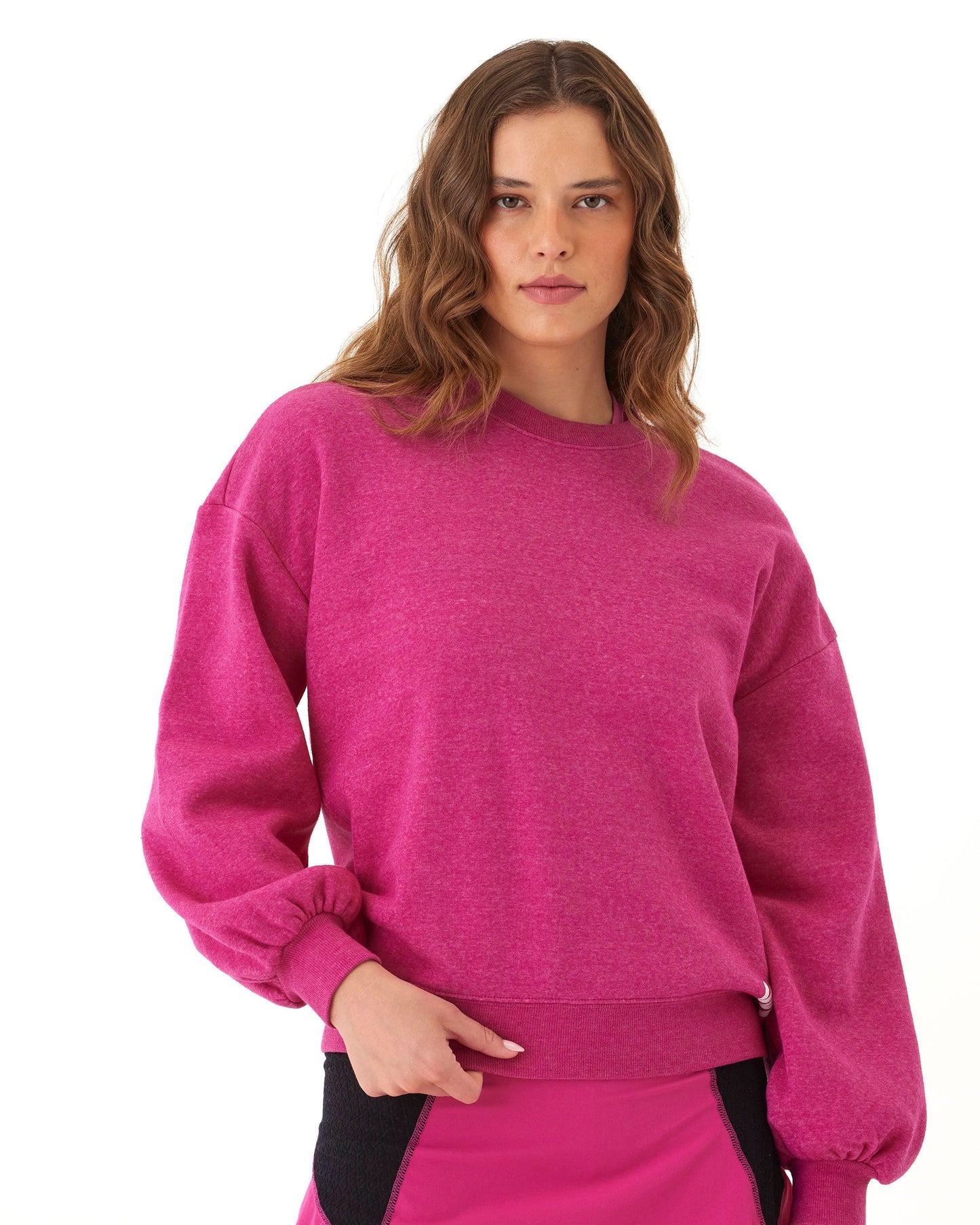 A woman with wavy brown hair stands against a white background, wearing Wringer Wear's bright pink Women's Crew Neck Sweatshirt and a matching pink skirt with black panels, giving her an athletic vibe. She looks at the camera with her hand on her waist.