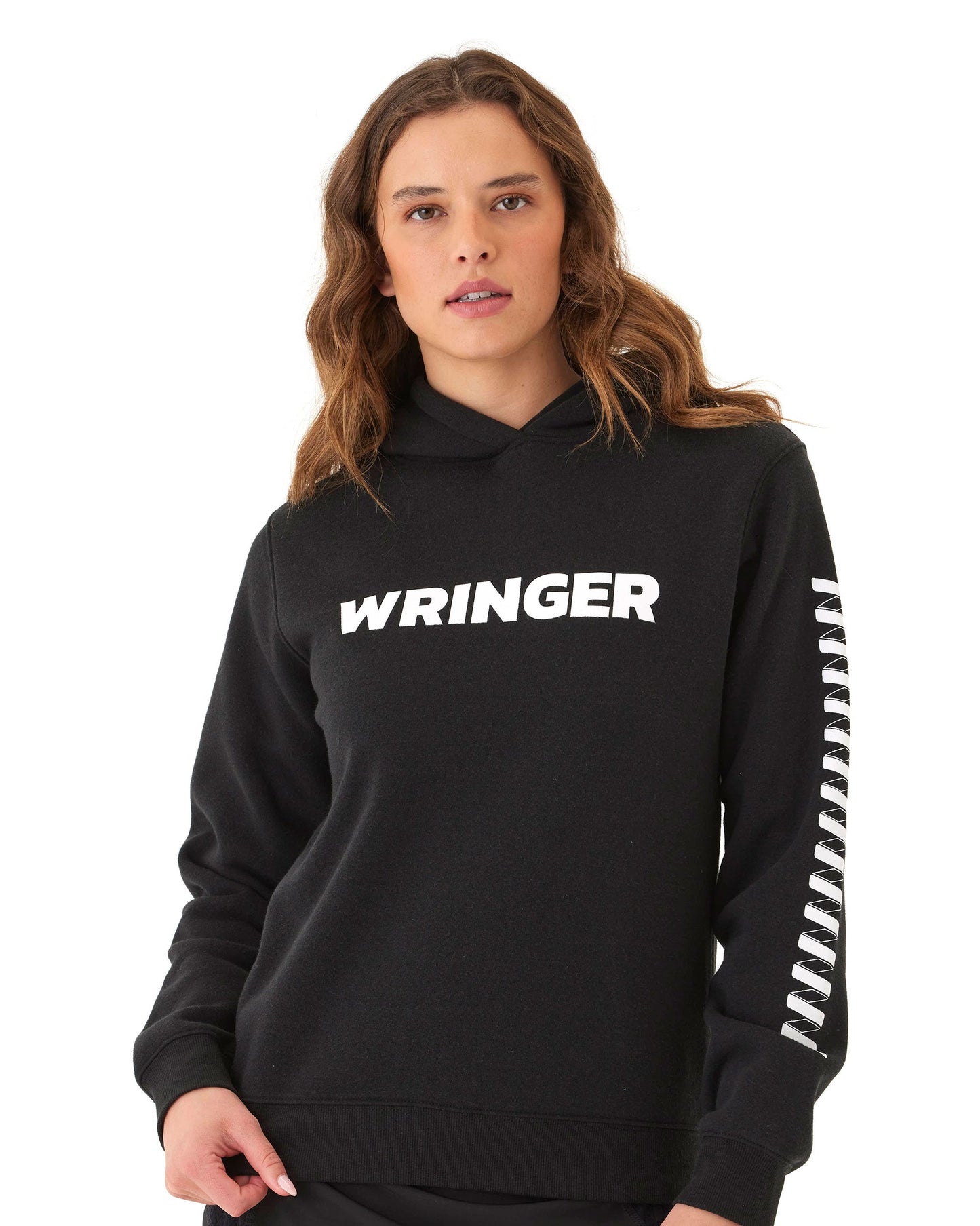 A young woman with wavy brown hair models the Women's Core Wringer Graphic Hoodie from Wringer Wear, featuring “WRINGER” in bold on the chest and a zigzag sleeve graphic. She stands against a white background, looking directly at the camera.
