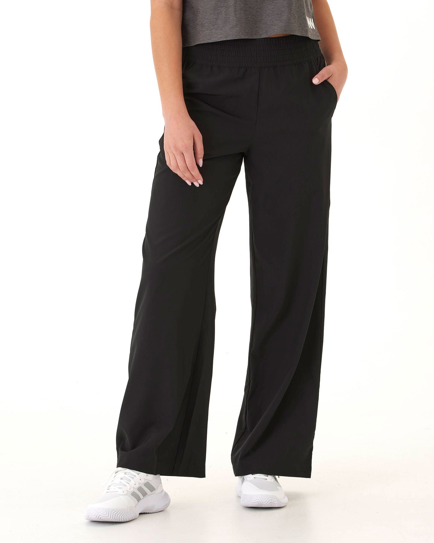 Against a plain white background, a person wears Wringer Wear Women's Wide Leg Pant made from hydrophobic stretch-woven fabric, styled with a gray top and white sneakers with gray stripes. Shown from shoulders down, one hand is in their pocket.