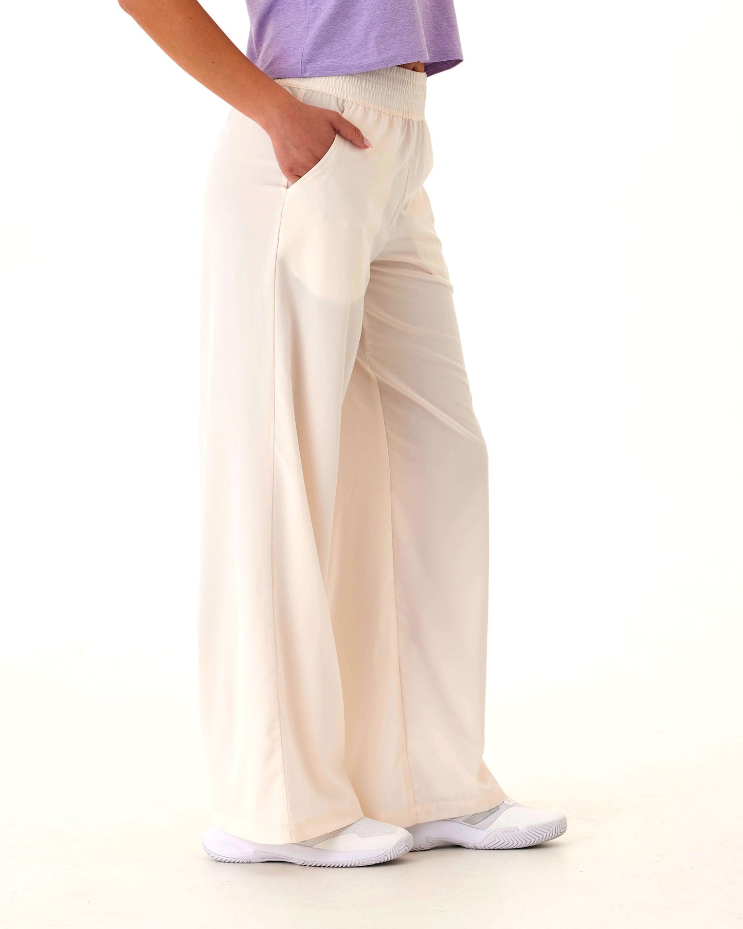 A person, lower half visible against a white background, models Wringer Wear’s Women's Wide Leg Pant in cream, made from hydrophobic stretch-woven fabric, paired with a light purple top and white slip-on shoes. Left hand is in the pocket.