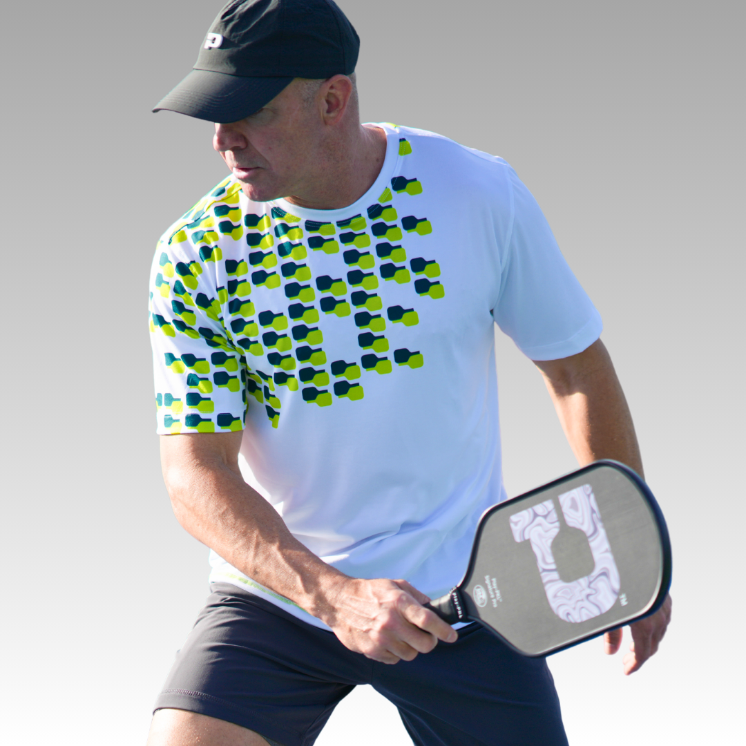 A man in the Playly Store’s Warrior Tee 2.0—a white pickleball shirt with green and blue patterns—plus black shorts and a cap, stands ready to play pickleball, holding a paddle with both hands against a plain gray background.