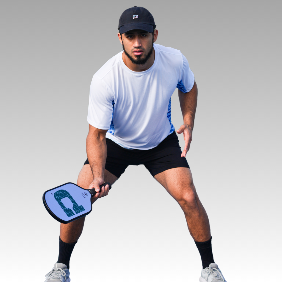 A young man in the Playly Store’s Warrior Tee 2.0, black shorts, and a black cap holds a pickleball paddle in a ready stance—slightly crouched with knees bent and eyes forward against a neutral gradient backdrop.