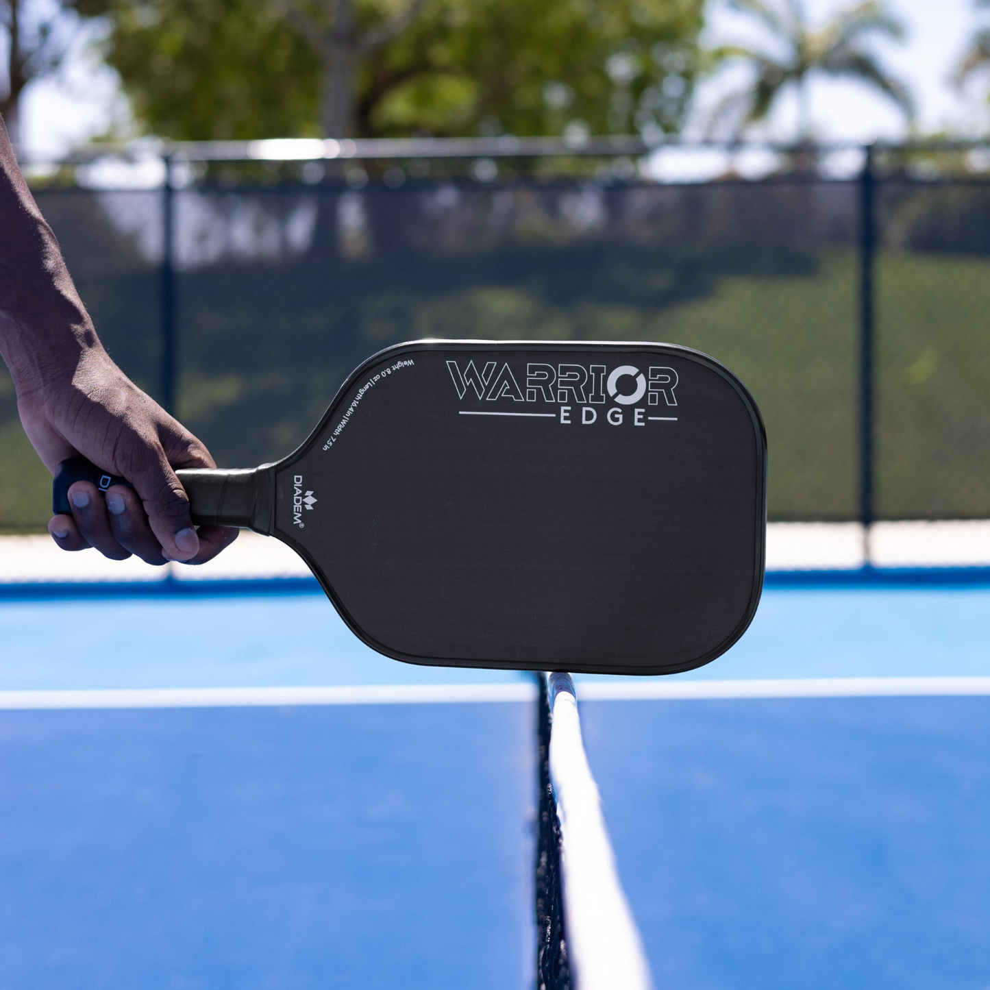 A person holds the Diadem Sports Warrior Edge paddle, featuring an etched carbon face, over a tennis net; this USAPA-approved design highlights the paddle's advanced craftsmanship.