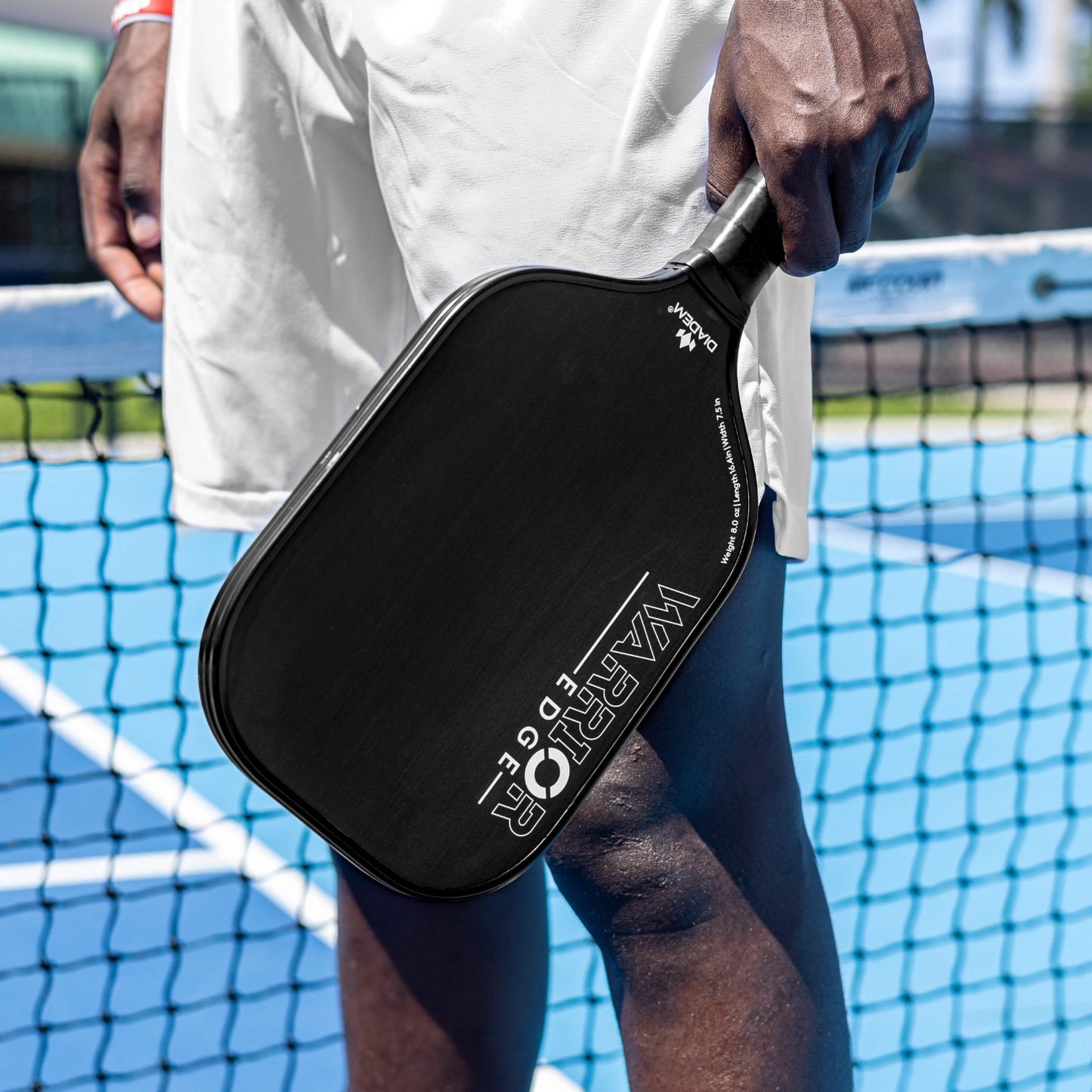A person in white shorts stands on a blue pickleball court, holding a black Diadem Sports Warrior Edge paddle by their side. A net stretches across the background as sunlight casts shadows on the court.