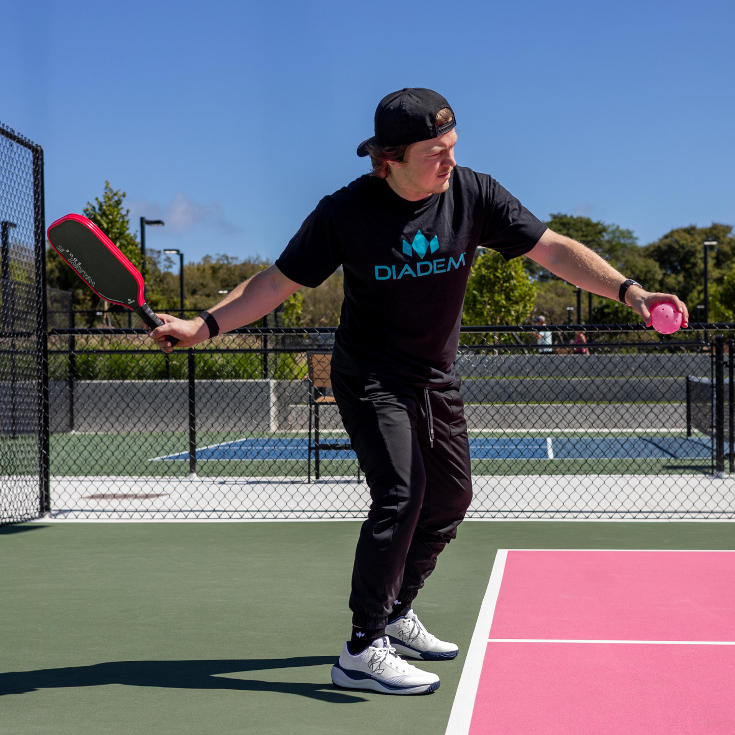 On a pink court, a player in black athletic wear and a backwards cap prepares to serve with the Diadem Sports Warrior Edge pickleball paddle. A blue tennis court and trees are seen in the background.
