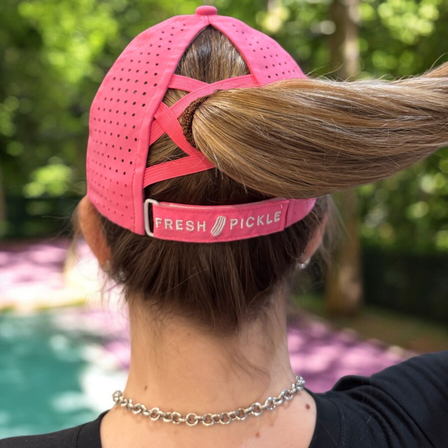 A person wears a pink Pickleball Cap by Fresh Pickle, with a ponytail through the back opening. The breathable sports hat features perforated holes and a “FRESH PICKLE” strap, set against blurred greenery and purple pavement.