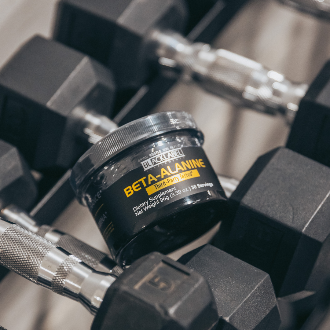 A BLACKLABEL Supplements BETA-ALANINE jar, labeled as a dietary supplement with 50g net weight and 52–70 servings, sits among hexagonal dumbbells in a gym, highlighting its third-party tested fitness focus.