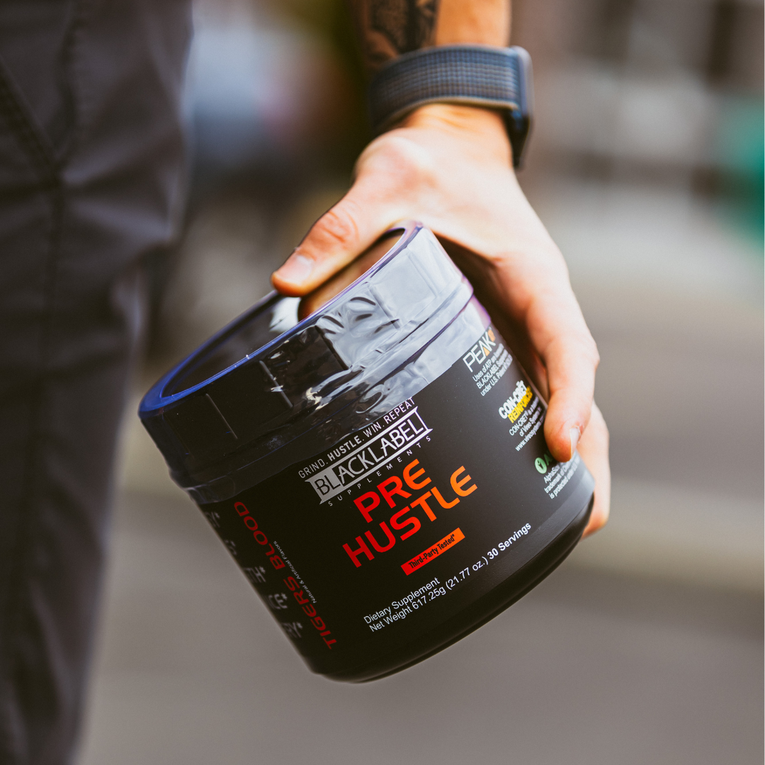 A hand wearing a blue fitness tracker holds a black container of PRE HUSTLE by BLACKLABEL Supplements, a pre-workout dietary supplement powder. The label highlights performance nutrients. The background is blurred.