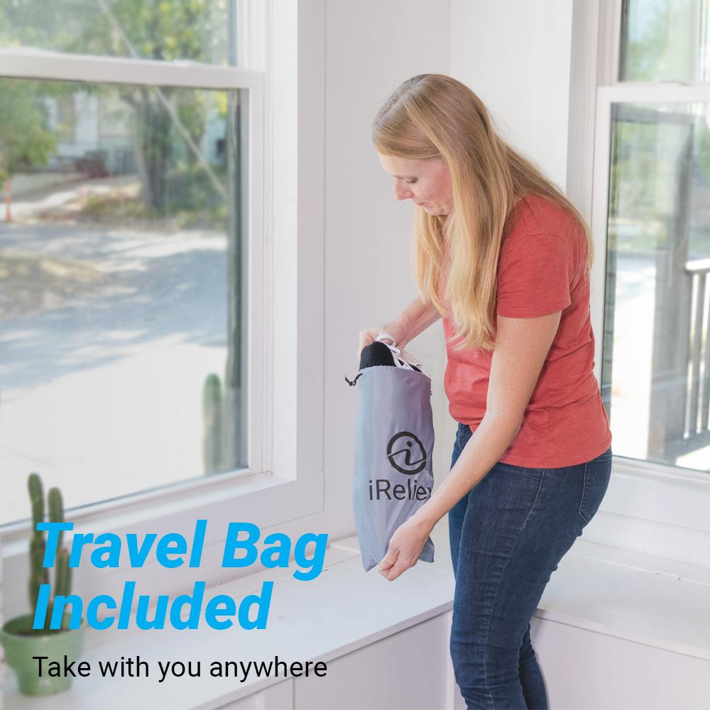 A woman with long blonde hair, in a red T-shirt and blue jeans, sits on a white windowsill holding a gray iReliev Weighted Heating Pad travel bag. Text reads: "Travel Bag Included. Take your iReliev Weighted Heating Pad anywhere." Sunlit windows behind her.