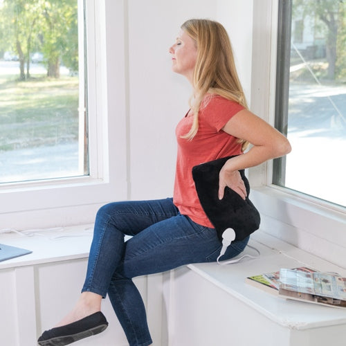 A woman in a red shirt and jeans sits on a windowsill, holding the iReliev Weighted Heating Pad to her lower back. Relaxed, she gazes outside. Nearby are a laptop, magazines, and trees seen through large windows.