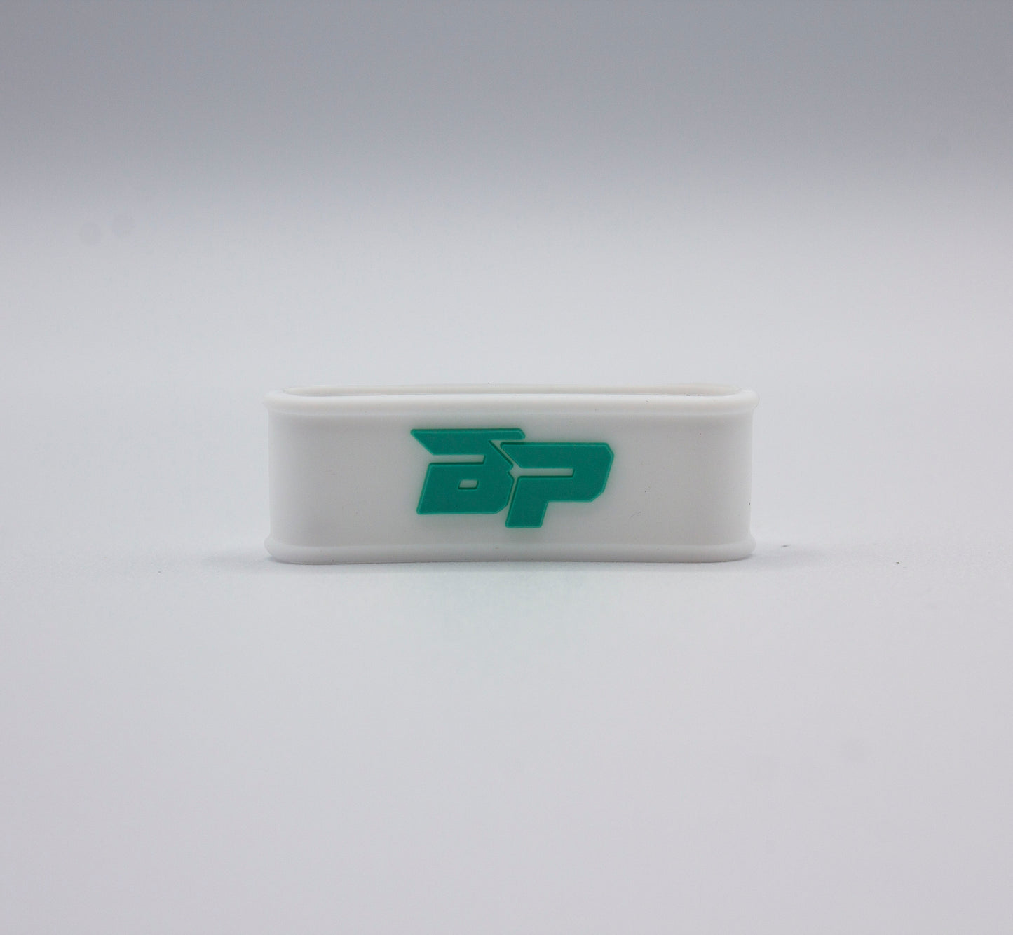 A small, rectangular white plastic Bodhi Performance Paddle Band with rounded edges stands upright. It features a bold teal BP logo on the front, resembling a pickleball paddle grip band against a minimalist white background.