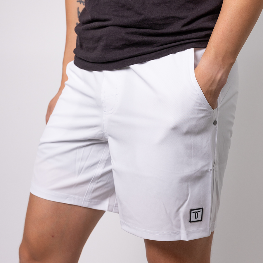 A person stands against a plain white background wearing black Thrive Pickleball Thrive Elite Shorts (7 in.) featuring a small black logo on the left leg, with one hand in a pocket and the other arm relaxed at their side.