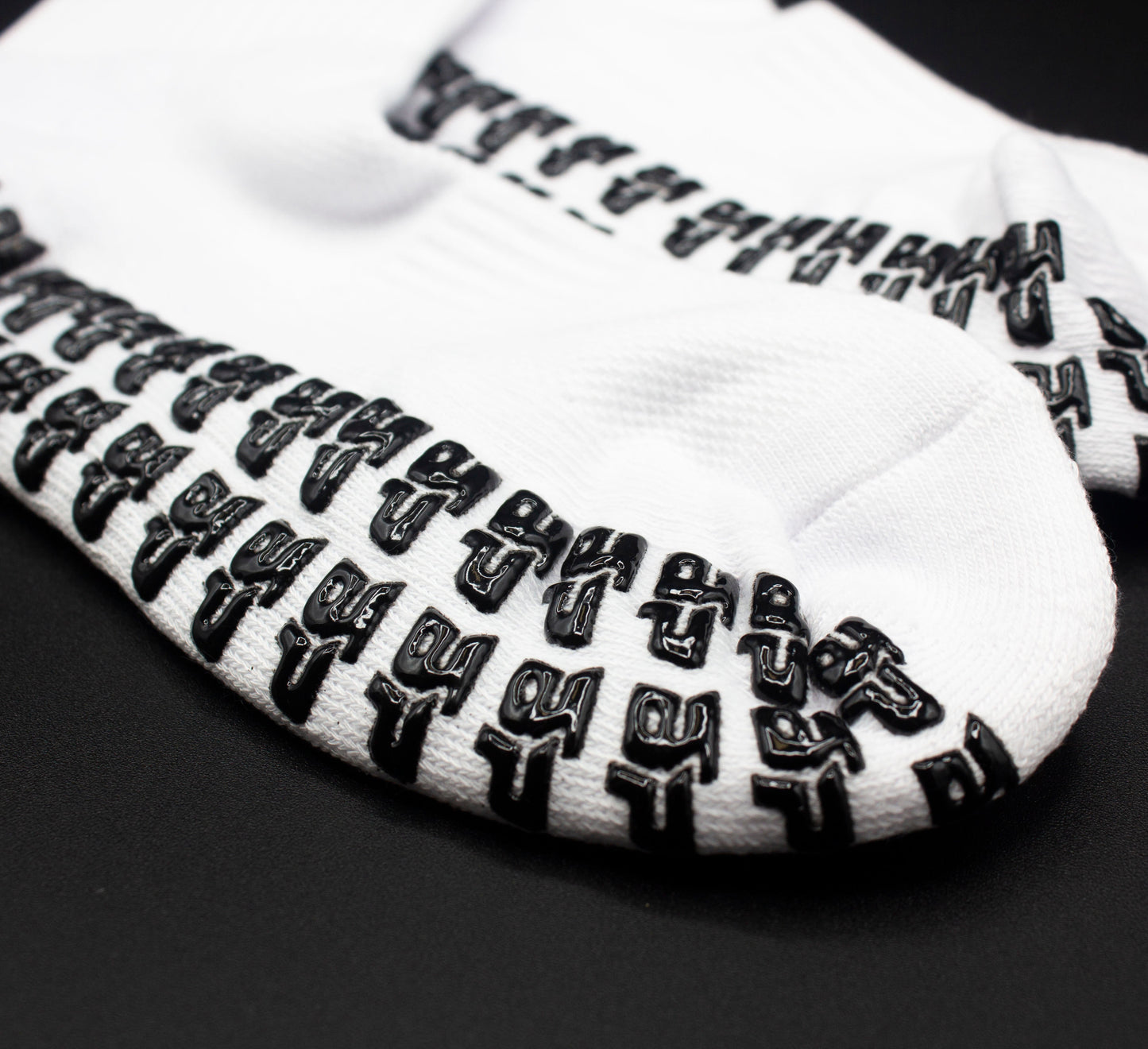 Close-up of Bodhi Performance's Performance Socks - GRIPtech™ in white, featuring black silicone grip patterns evenly spaced on the soles for traction—ideal for yoga, pilates, and other activities. Arranged on a black surface.