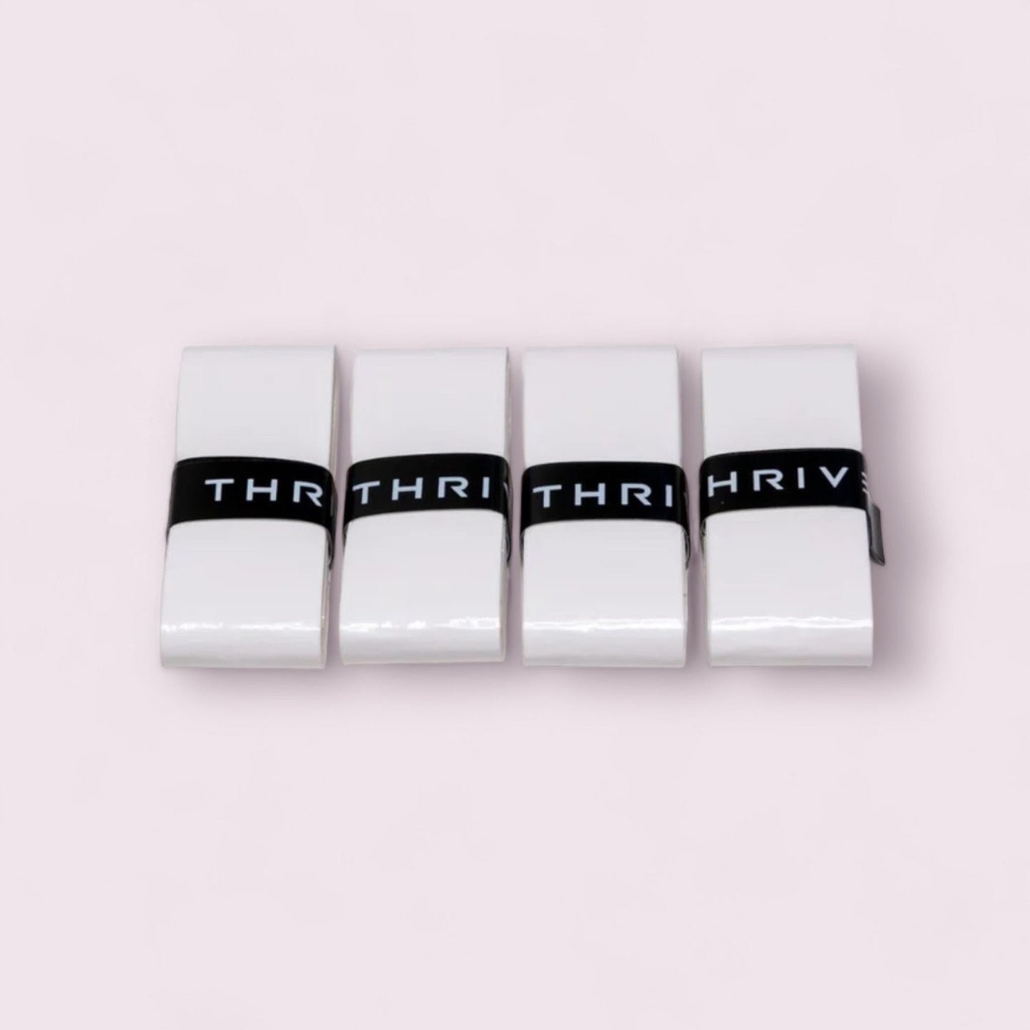 Four rectangular white Thrive Pickleball Overgrips (Super Tacky) are stacked side by side on a light pink background. Each is wrapped with a black band featuring bold white “THRIV” lettering, highlighting durability and grip.