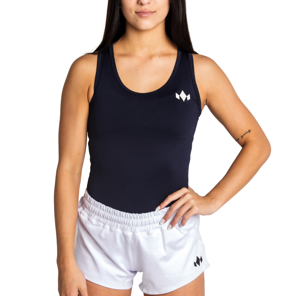 A woman with long dark hair and a tattoo on her left forearm poses with one hand on her hip, wearing a black Diadem Sports Womens Essential Drycore Athletic Moisture-Wicking Training Top and white shorts. The background is plain white.