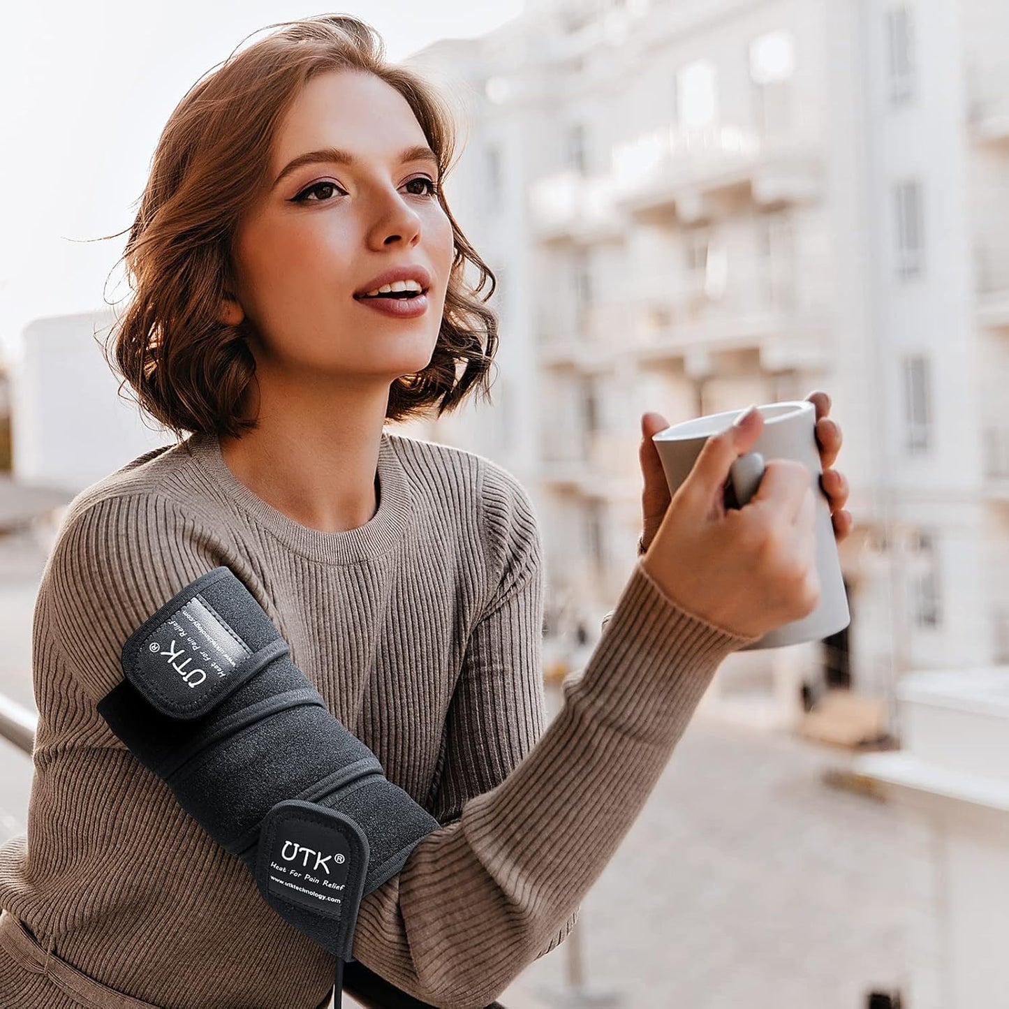 A woman with light brown hair and a beige ribbed sweater sits outside, smiling and holding a white mug. An iReliev Far Infrared Heating Pad Wrap with Far Jade is wrapped around her right arm. A sunlit urban street is softly blurred in the background.