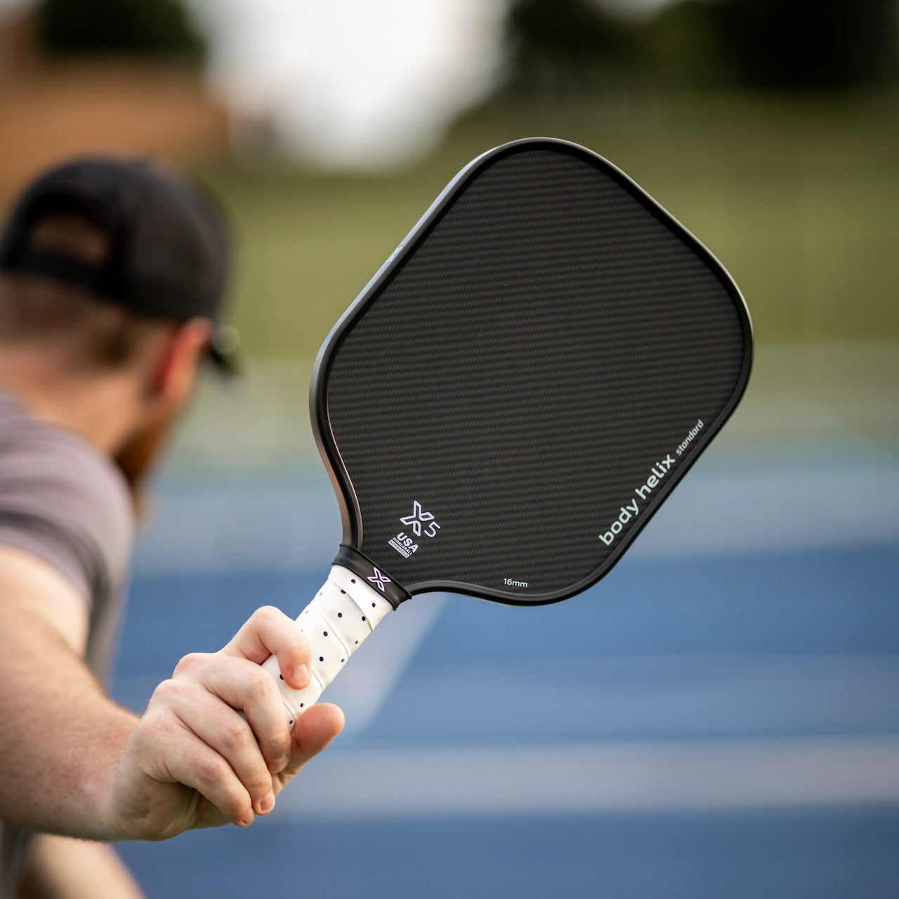 A person holds the body helix X5 Kevlar Pickleball Paddle—USAP Approved—with a cushioned grip and white handle, seen close-up from behind on an outdoor blue court, with blurred greenery hinting at an exciting game in progress.