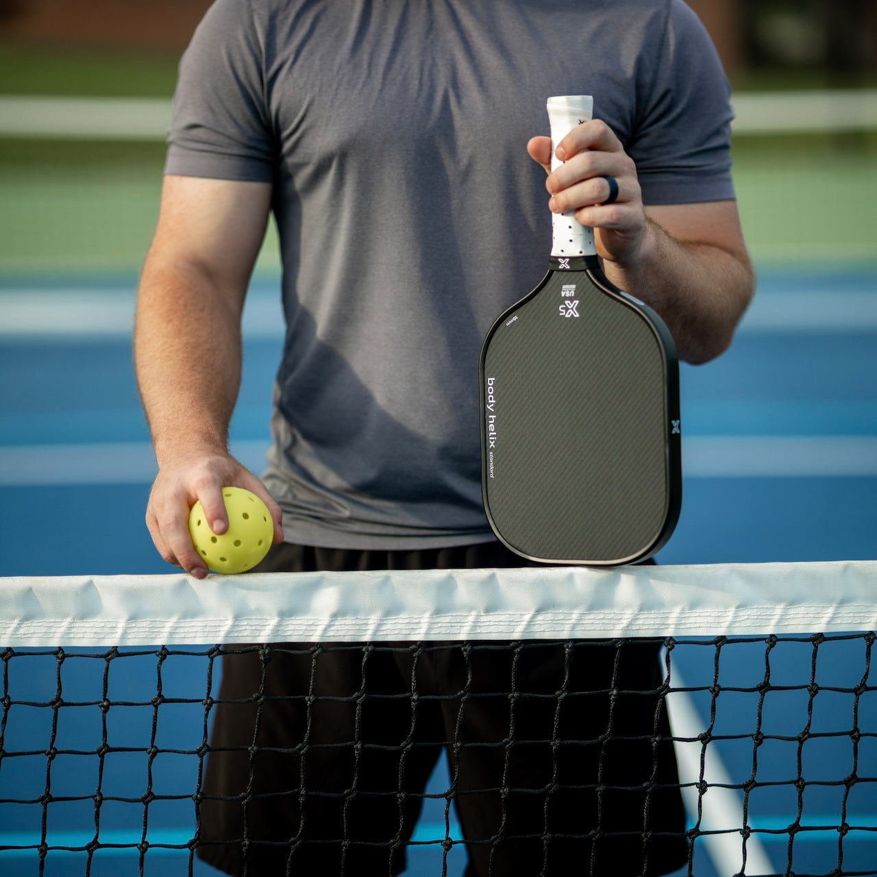 A person wearing a gray t-shirt and black shorts stands on a blue pickleball court, holding a yellow pickleball and the body helix X5 Kevlar Pickleball Paddle - USAP Approved with a cushioned grip, near the net. Only their torso and arms are visible.