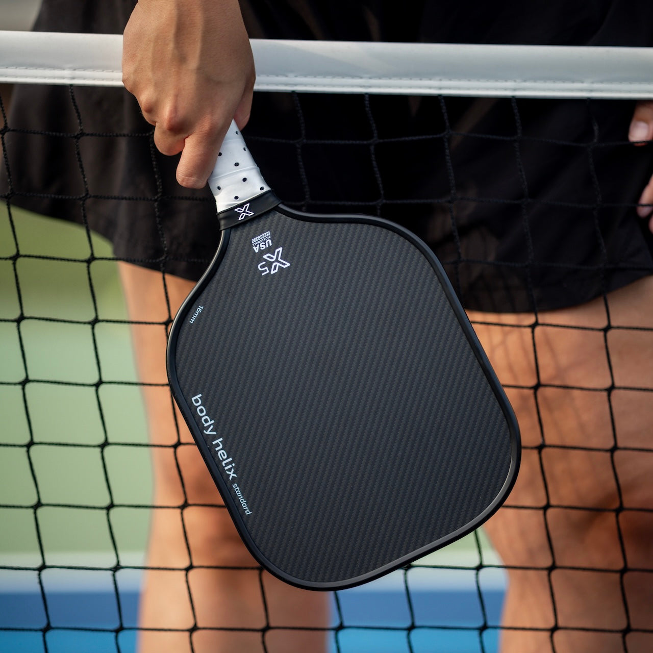 A person holds the body helix X5 Kevlar Pickleball Paddle - USAP Approved, featuring a cushioned grip and sleek, textured black finish, over a net on an outdoor court. Only their hand, part of their shorts, and the paddle are visible.