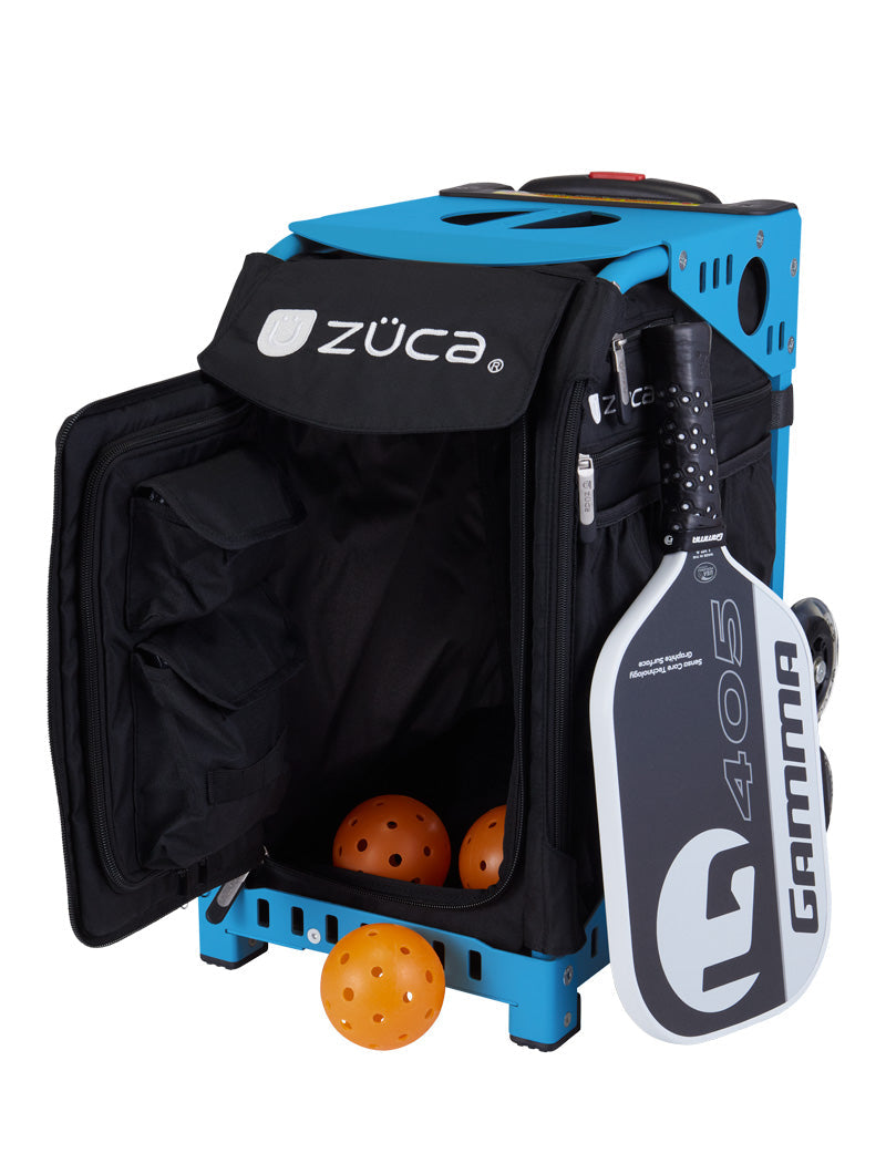 A ZUCA Pickleball Sport Black/Black bag sits with its front compartment open, showing three orange pickleballs inside. A black-and-white GAMMA G405 paddle is attached outside. The rolling bag features side pockets and metal frame details for convenience.