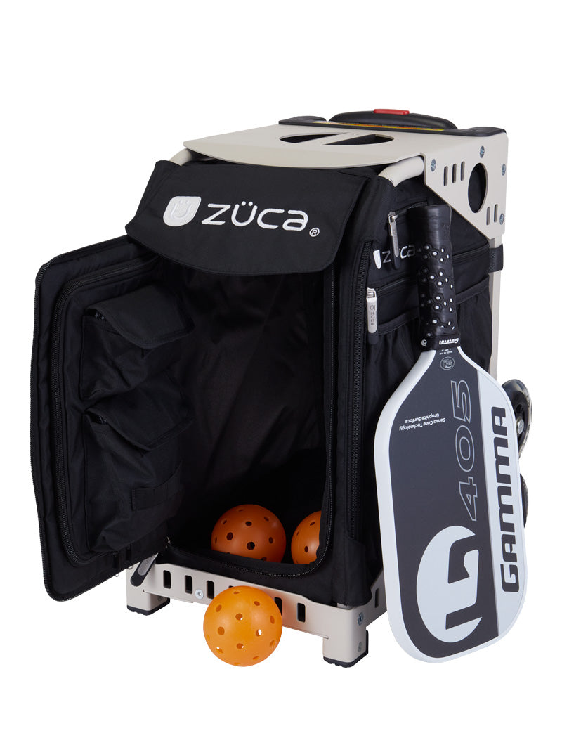 The ZUCA Pickleball Sport Black/Black bag has wheels for easy transport and a side door revealing three orange pickleballs inside, with a black and white Gamma paddle attached outside. This sturdy, spacious bag is perfect for pickleball storage.