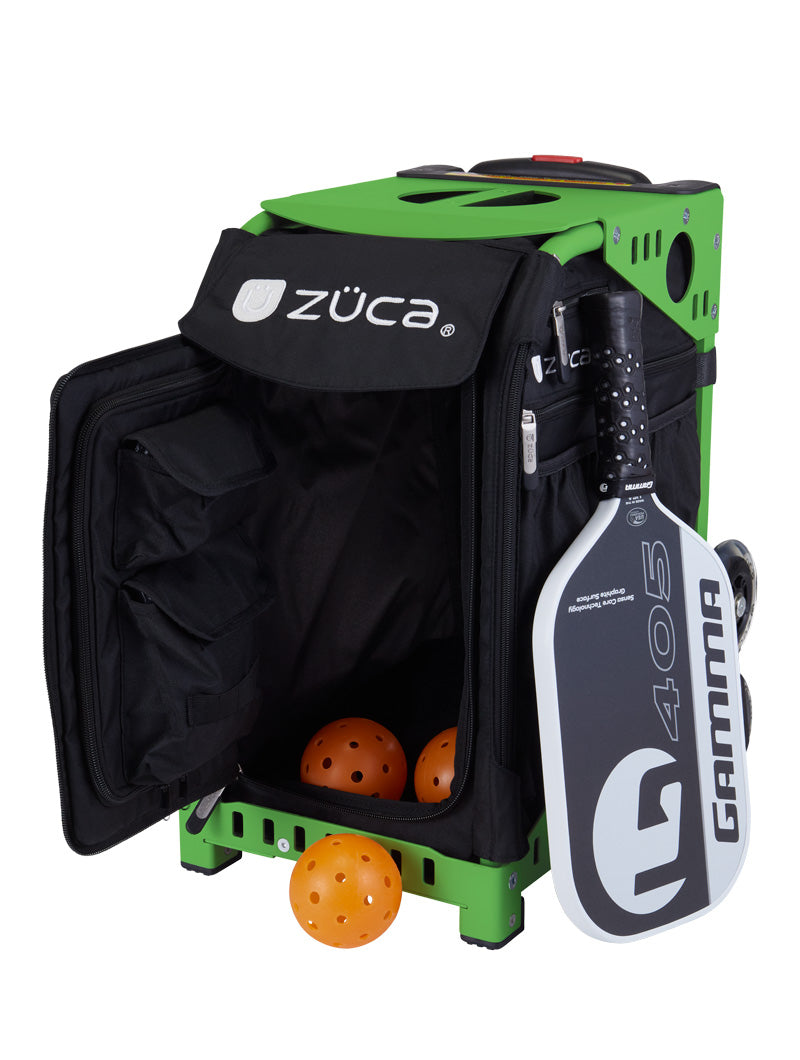 A ZUCA Pickleball Sport Black/Black rolling bag is open, revealing three orange balls inside. A black and white Gamma paddle hangs on the side. This wheeled storage bag includes a black interior and convenient side pockets.