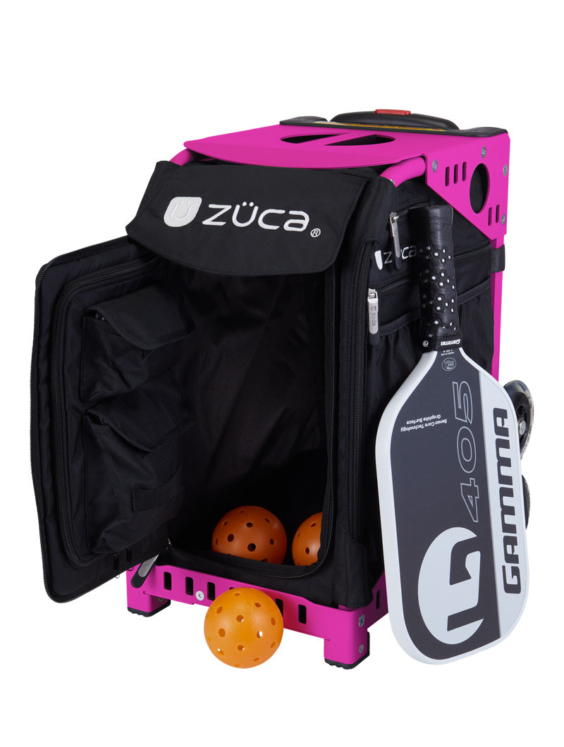 A ZUCA Pickleball Sport Black/Black rolling bag with its front pocket open reveals yellow pickleballs inside. A white and black Gamma 405 paddle is attached to the side. The bag features sturdy wheels, black handle straps, and ample storage.