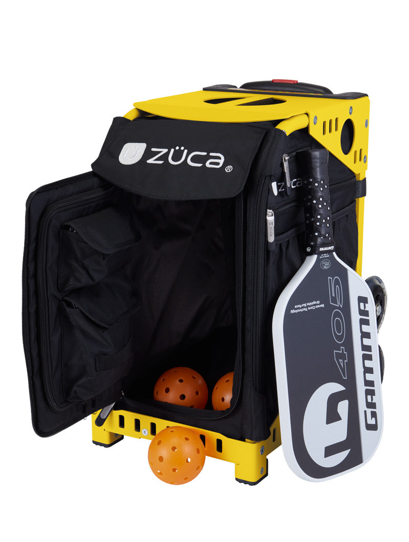 A ZUCA Pickleball Sport Black/Black rolling bag with an open front compartment displays three orange perforated pickleballs. A black-and-white Gamma 405 paddle is attached to the outside. The wheeled bag offers side pockets and ample space for gear.