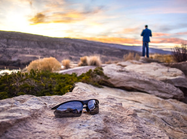 A pair of Cadence Lightweight Wrap Sunglasses by Epoch Eyewear with UV protection rests on a sunlit rock, while in the background, someone admires a colorful sunset over hills and a river beneath soft clouds. The atmosphere is calm and scenic.