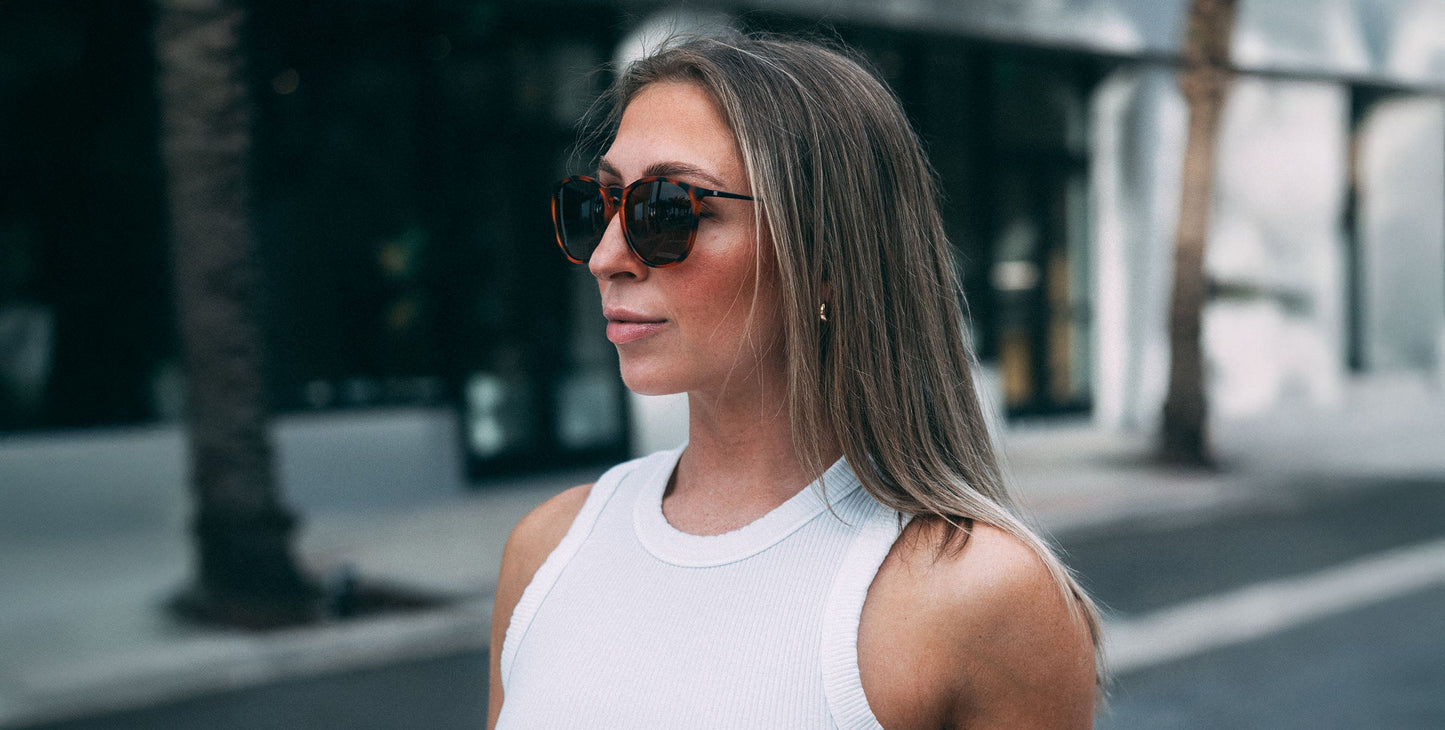 A woman with straight, light brown hair wears Neven Eyewear’s Bowie sunglasses—large tortoise frames with polarized UV400 lenses—paired with a white sleeveless top, standing confidently on a city street with palm trees in the background.