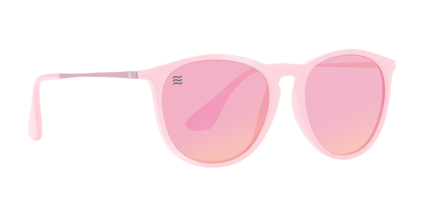 The Dreamy by Neven Eyewear features round, light pink plastic frames with matching pink-tinted, impact-resistant lenses, thin silver and pink temples, and a small wavy logo on the top left lens. Photographed on a plain white background.