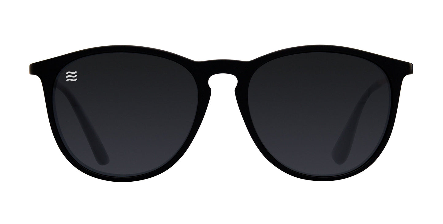 The Goldie Lux by Neven Eyewear are black and gold sunglasses with round, dark-tinted lenses, a slight cat-eye shape, TR90 frames, and a small white wavy logo on the left lens. Photo features a plain white background.