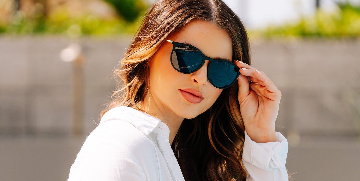 A woman with long, wavy brown hair wears Neven Eyewear’s Goldie Lux sunglasses in black and gold with polarized UV400 protection. Dressed in a white shirt, she lifts her shades while looking at the camera outdoors.