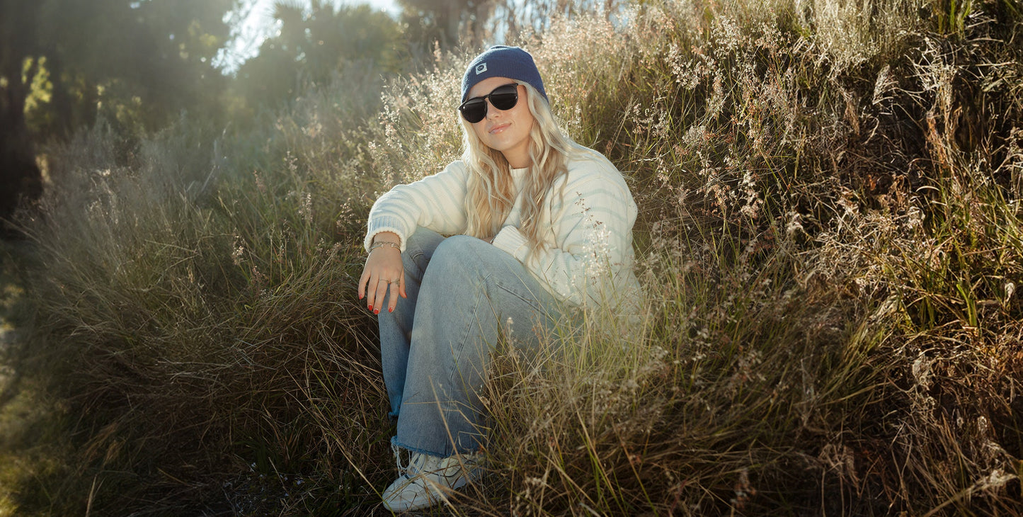 A woman with long blonde hair sits in tall grass, wearing Neven Eyewear’s Goldie Lux sunglasses with polarized UV400 protection, a blue cap, cream sweater, and loose blue jeans. One knee up, she enjoys the sunlight and greenery.