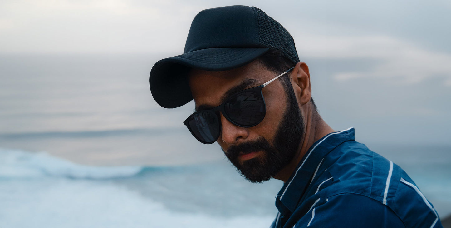 A man with a trimmed beard and mustache wears Neven Eyewear’s Goldie Lux dark sunglasses with polarized UV400 protection, a black baseball cap, and a blue jacket with white stripes. He faces the camera, serious, against cloudy sky and ocean waves.