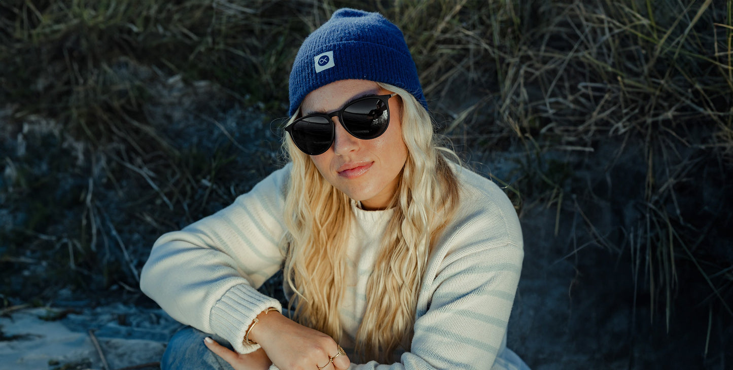 A person with long blonde wavy hair wears Neven Eyewear's Goldie Lux sunglasses and a blue knit beanie, paired with a cream striped sweater, sitting outdoors in front of tall grass and sand, looking at the camera with a relaxed expression.