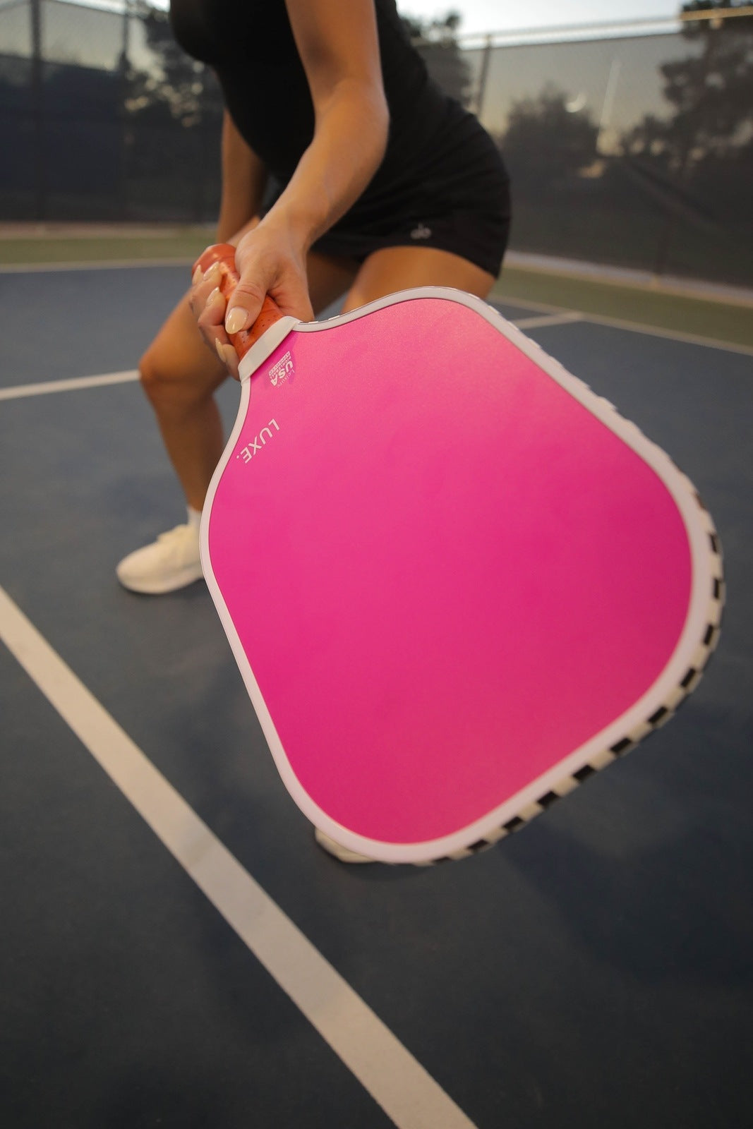On a blue pickleball court, a person in black shorts, black top, and white sneakers holds the LUXE Pickleball Aesthetic Solids - Leisure pink fiberglass paddle toward the camera. Only their lower body is visible. A chain-link fence and trees are in the background.