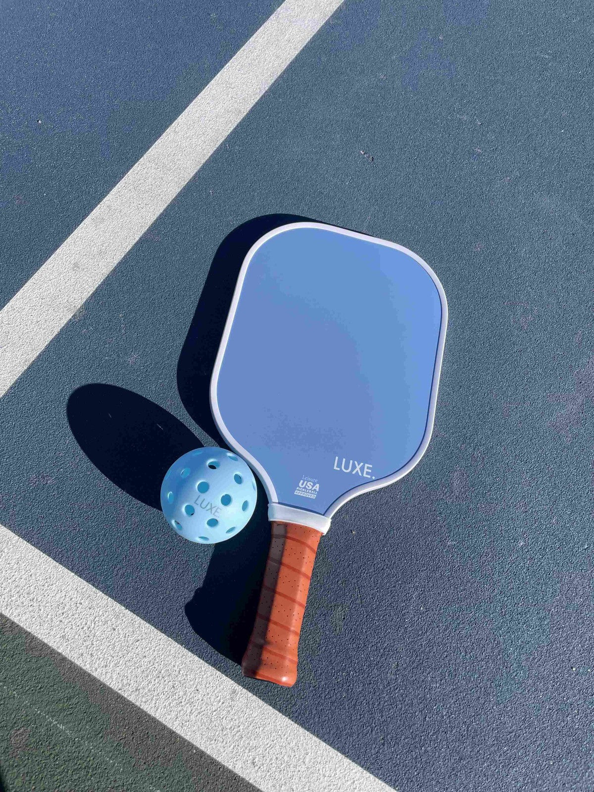 A blue Aesthetic Solids - Leisure fiberglass paddle by LUXE Pickleball, USA Pickleball Approved, with a brown handle and blue perforated ball, rests on a blue-green court. The “LUXE” paddle casts leftward shadows over the white lines.