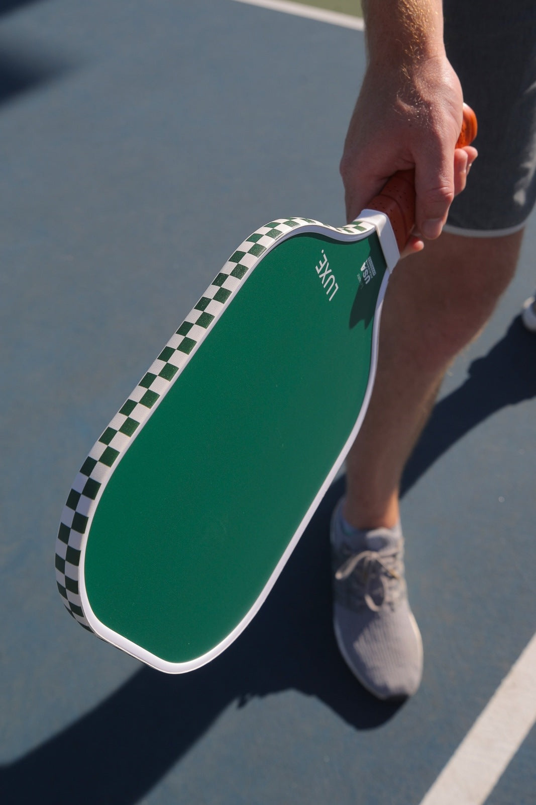 A person holds the LUXE Pickleball Aesthetic Solids - Leisure fiberglass paddle with a honeycomb core and white-green checkered edge on a blue court. Only their lower body in gray shorts and sneakers is visible. USA Pickleball Approved.