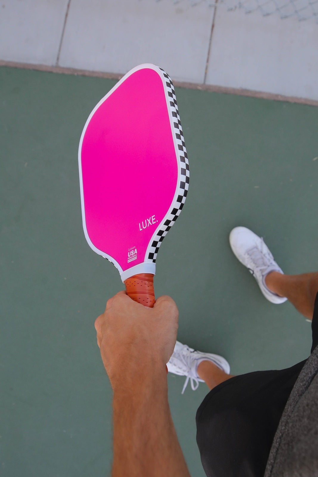 A person holds the bright pink "Aesthetic Solids - Leisure" fiberglass paddle by LUXE Pickleball, with a black-and-white checkered edge, honeycomb core, and light brown leather grip, on a green court. USA Pickleball Approved.