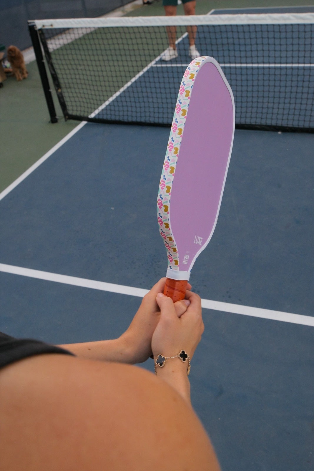 On an outdoor court, someone holds the LUXE Pickleball Aesthetic Solids - Leisure paddle—pastel purple fiberglass with a colorful edge and orange grip. Its honeycomb core is visible as another player and a dog stand near the net in the background.