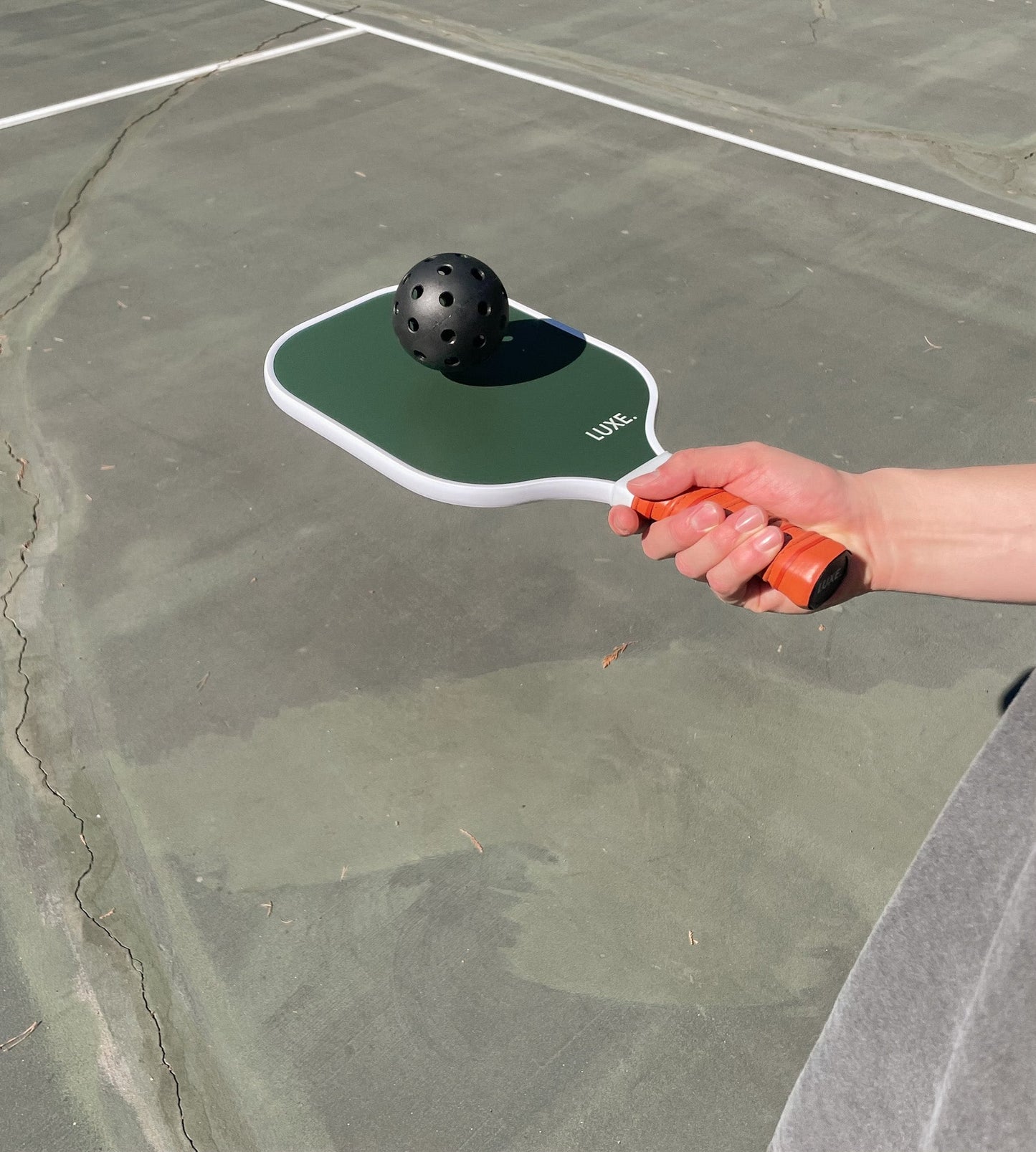 A person holds the LUXE Pickleball "Aesthetic Solids - Leisure" green and white fiberglass paddle with an orange grip, balancing a black pickleball on its honeycomb core while standing on a cracked outdoor court with white boundary lines.