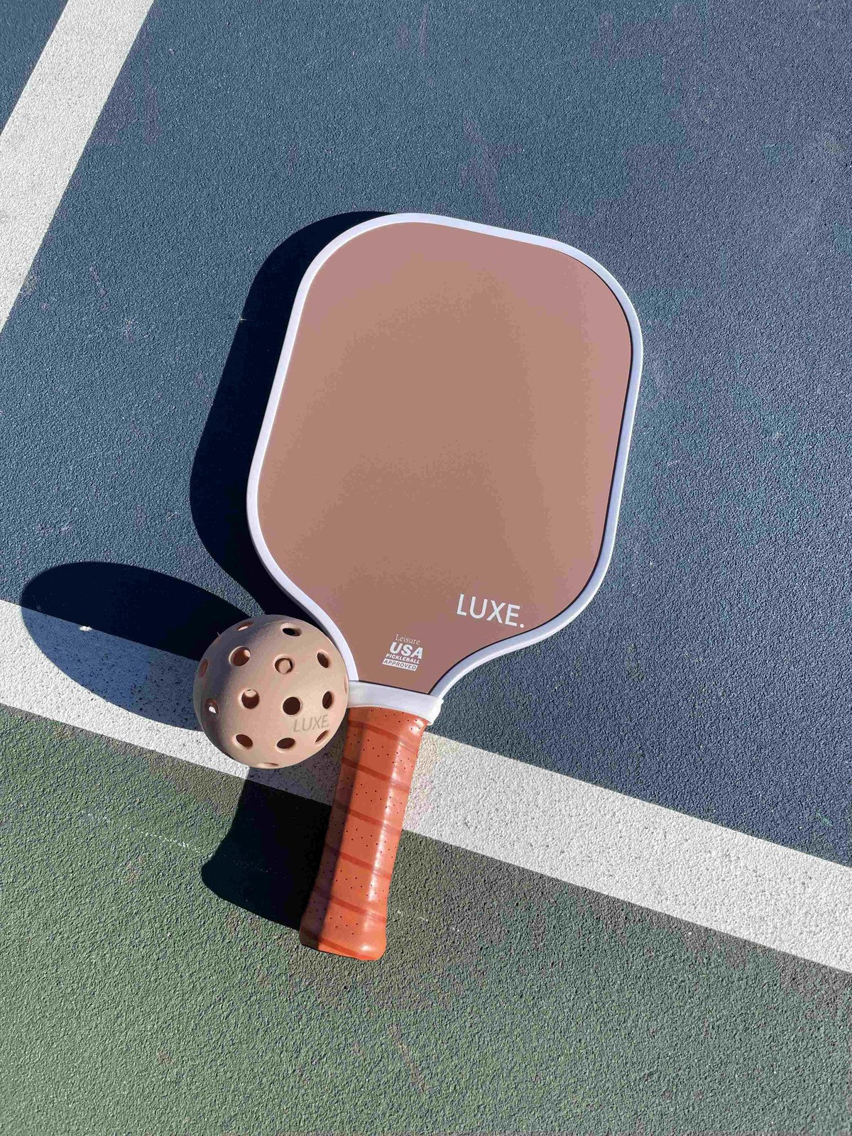 A LUXE Pickleball Aesthetic Solids - Leisure paddle with a tan leather grip rests on a vibrant court next to a tan pickleball. The fiberglass paddle’s honeycomb core and USA Pickleball approval gleam in the sunlight beside crisp white lines.