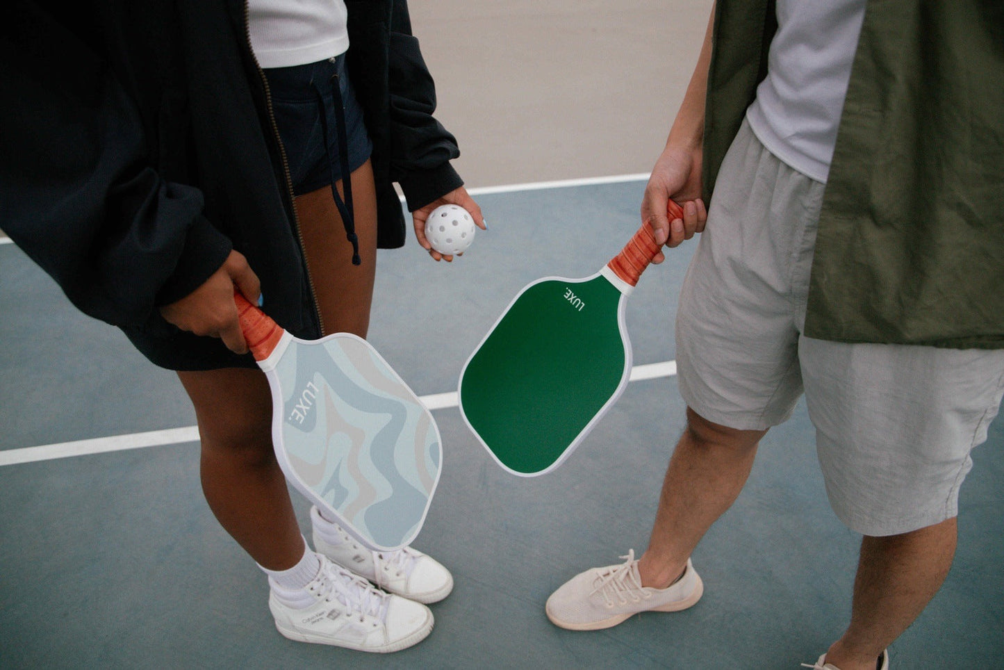 Two people on an outdoor court each hold a LUXE Pickleball Aesthetic Solids - Leisure paddle with a honeycomb core. The “JADE” and “JOE” paddles are USA Pickleball Approved. Both wear sporty casual outfits and sneakers.