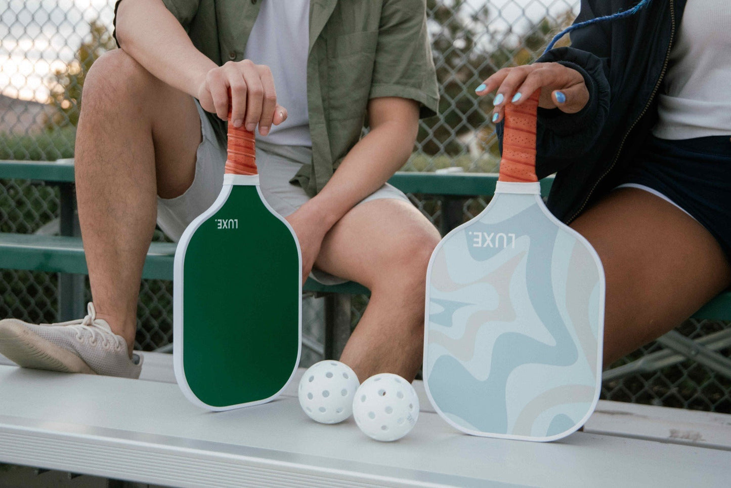 Two people sit on bleachers, each holding a LUXE Pickleball Aesthetic Solids - Leisure fiberglass paddle—one green, one white with blue designs and honeycomb core—both USA Pickleball Approved. Three perforated pickleballs rest between them by the chain-link fence.