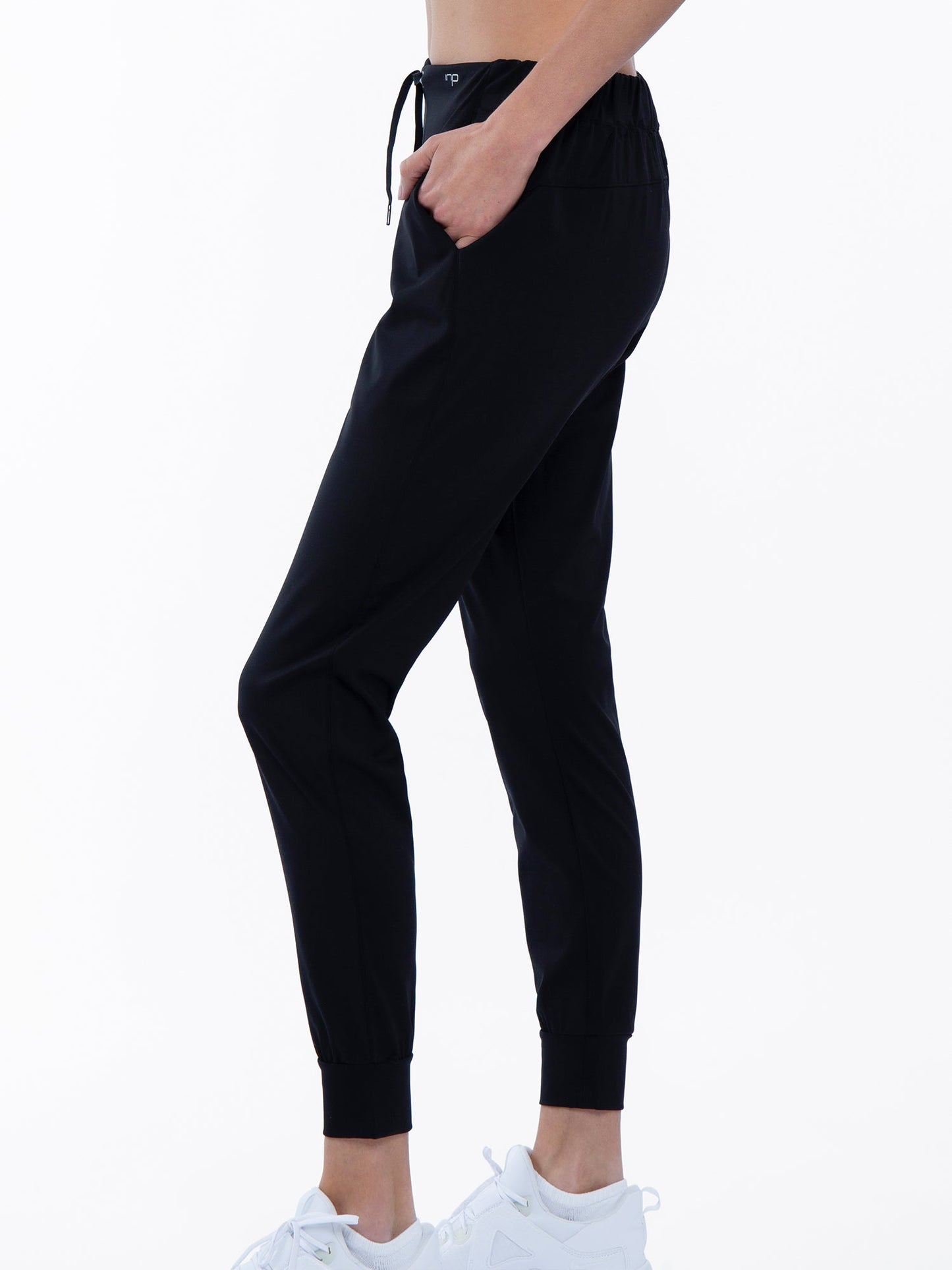 A person stands in profile wearing the inPhorm NYC Alexa Jogger in black, featuring an elastic waistband, cuffed ankles, and pockets. White sneakers and a plain white backdrop highlight the fit and style of these joggers.